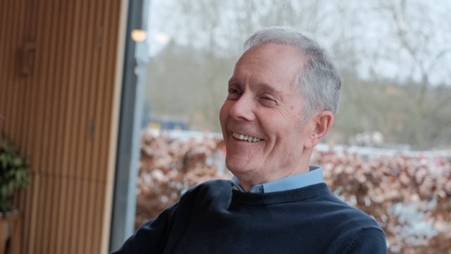 An older man with short gray hair and a light complexion is sitting indoors, smiling warmly. He is wearing a dark sweater over a light blue collared shirt. The background shows a window with an out-of-focus view of trees and foliage outside.